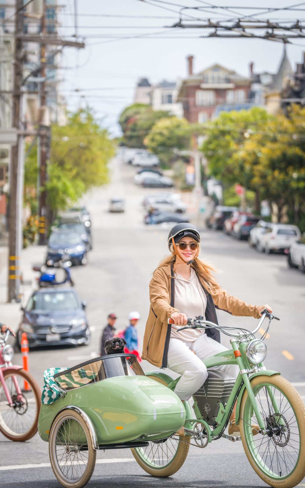 A woman riding a sage-green Mad Dogs & Englishmen electric bike with a sidecar carrying a small dog, on a San Francisco hill